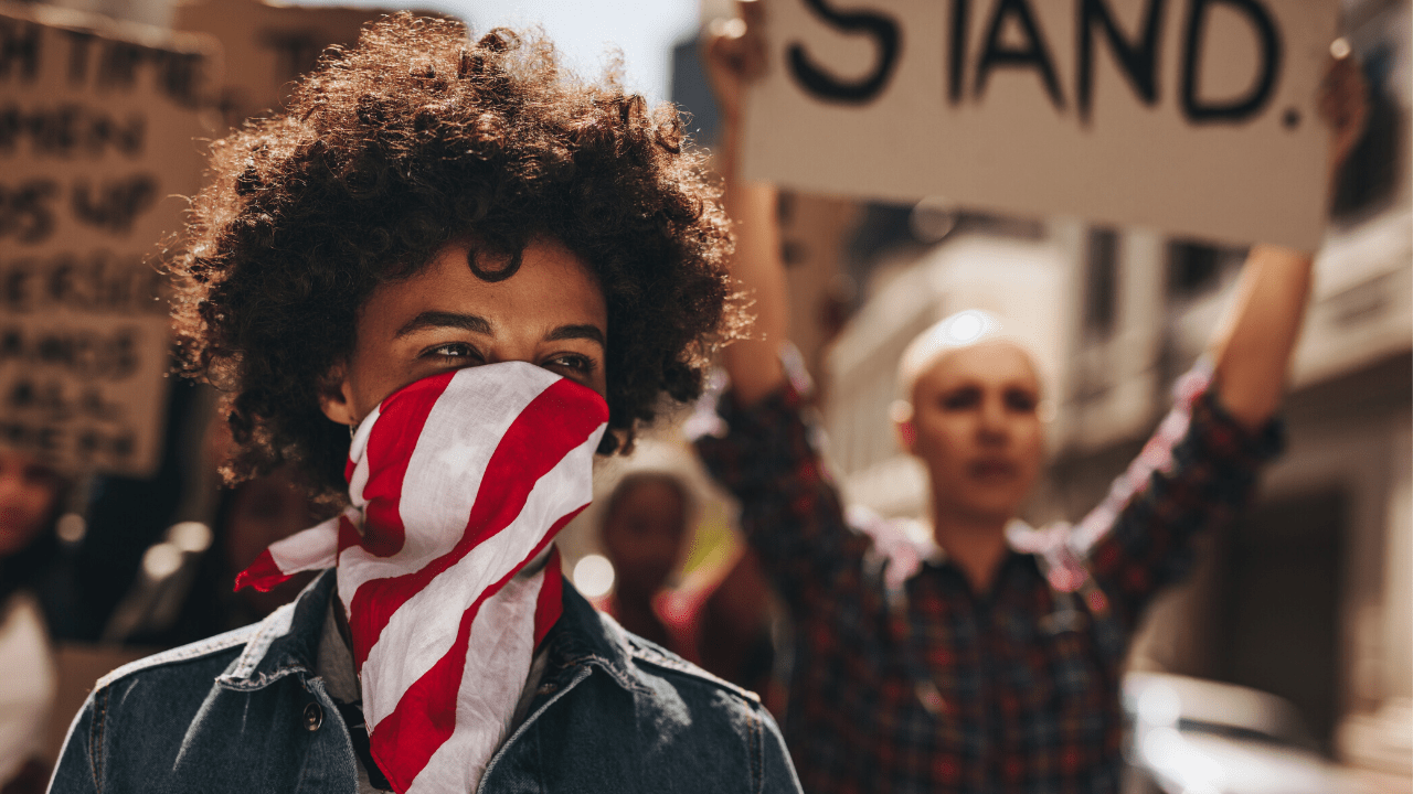 A Black woman with an american flag covering her mouth. Protesters stand behind her holding signs and marching.