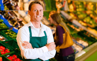 A light skinned male with brown hair working in a grocery store standing in front of the produce smiling.