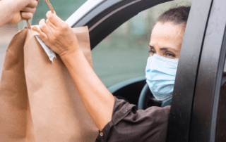 A female wearing a mask is sitting in her car with the window down and taking a brown bag that is being handed to her from outside the vehicle.