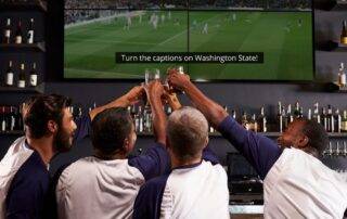 A group of sports fans in a bar. Their backs visible and holding up their drinks to the TV with captions displayed.