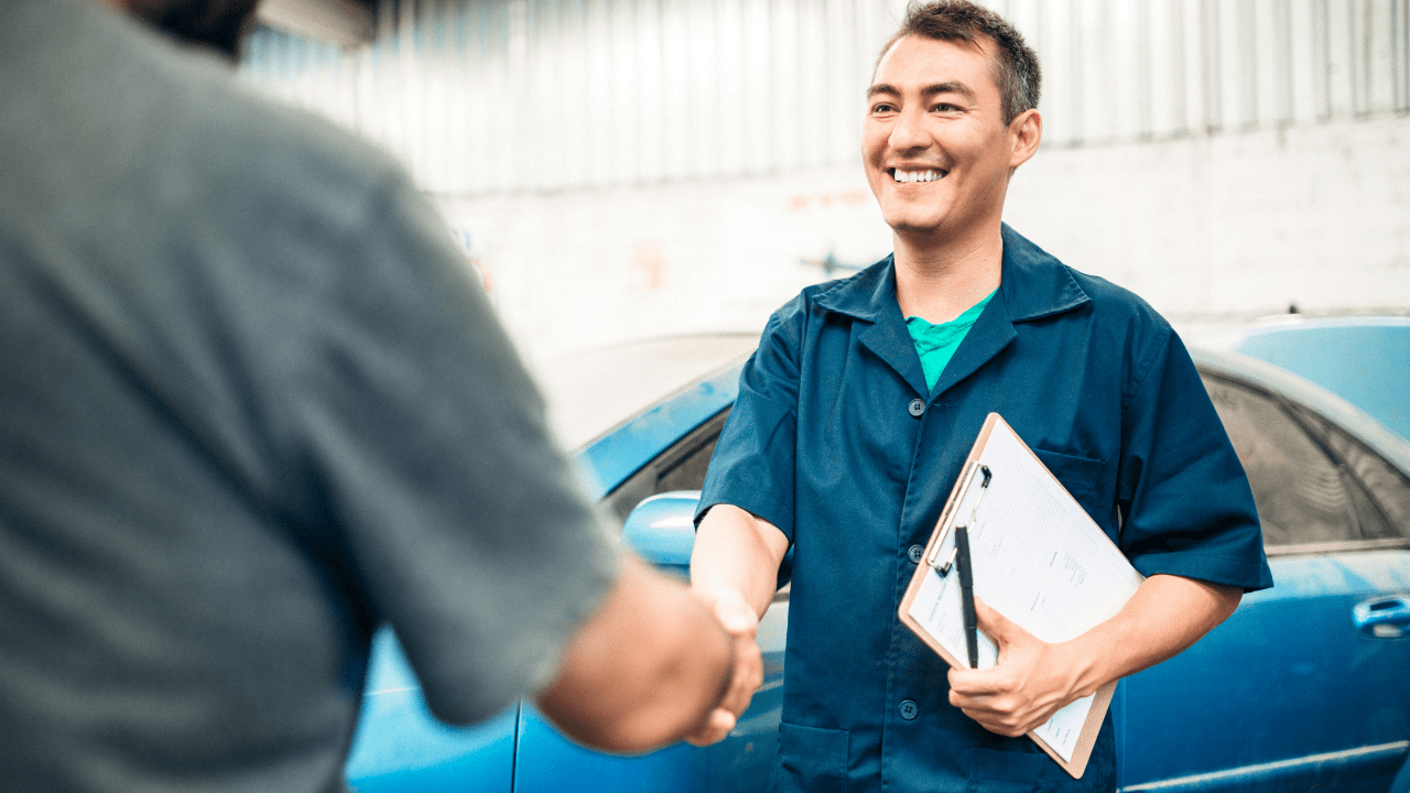 An auto mechanic holding a binder with a blue car in the background is shaking the hand of a customer.