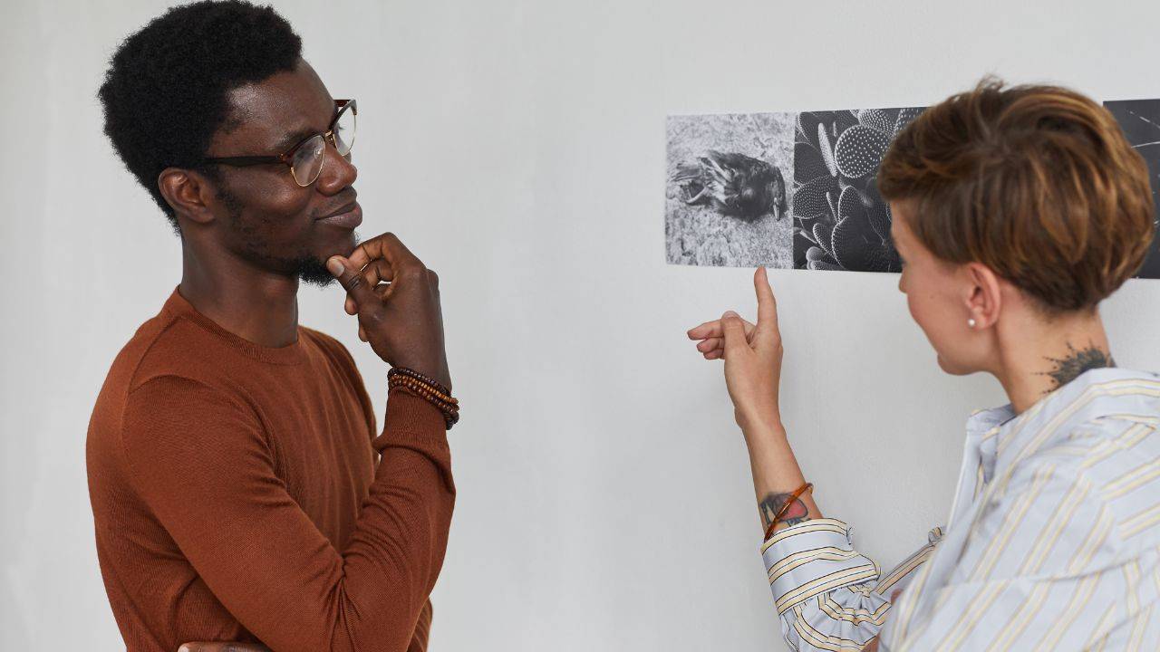 A fair skinned woman with short hair and a neck tattoo points to art on the wall. A black man with glasses has his hand on his chin and is focused on the woman and her explaining of the art on the wall.