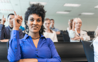A dark skinned female with her hair up learning sign language. She is wearing a blue top and is smiling. There are other students behind her using sign language out of focus.